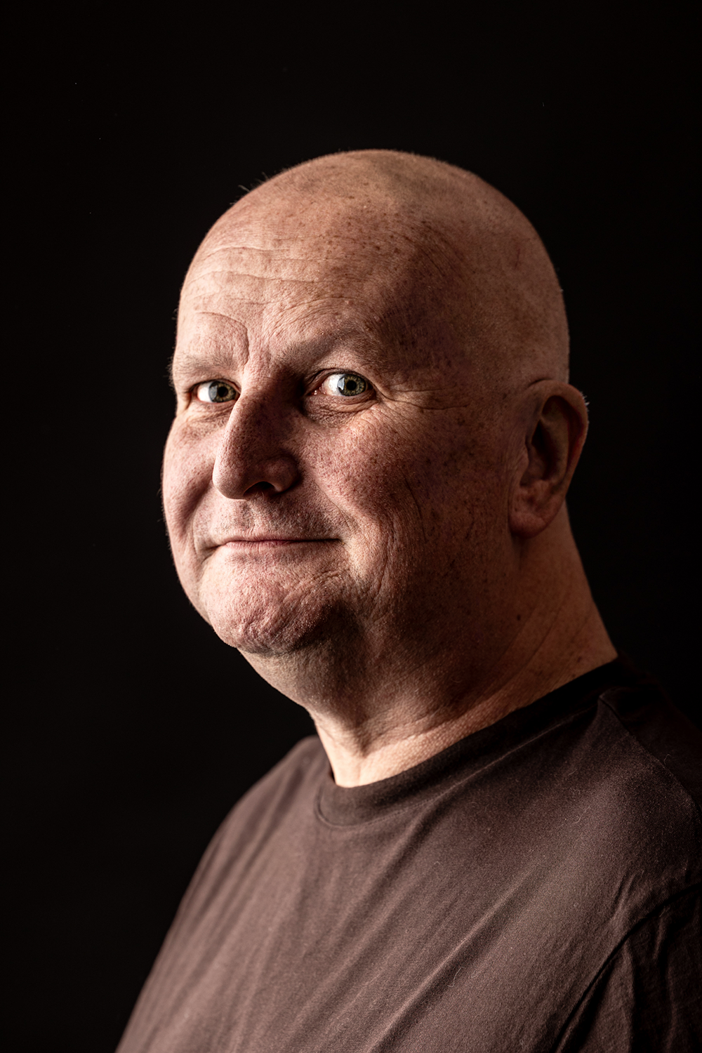 Portret dojrzałego, łysego mężczyzny w ciemnej koszulce, sfotografowanego w miękkim bocznym świetle na czarnym tle. Naturalny wyraz twarzy i subtelny uśmiech.